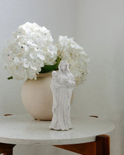 Statue of a religious figure with a child in a vase of white flowers on a marble table.