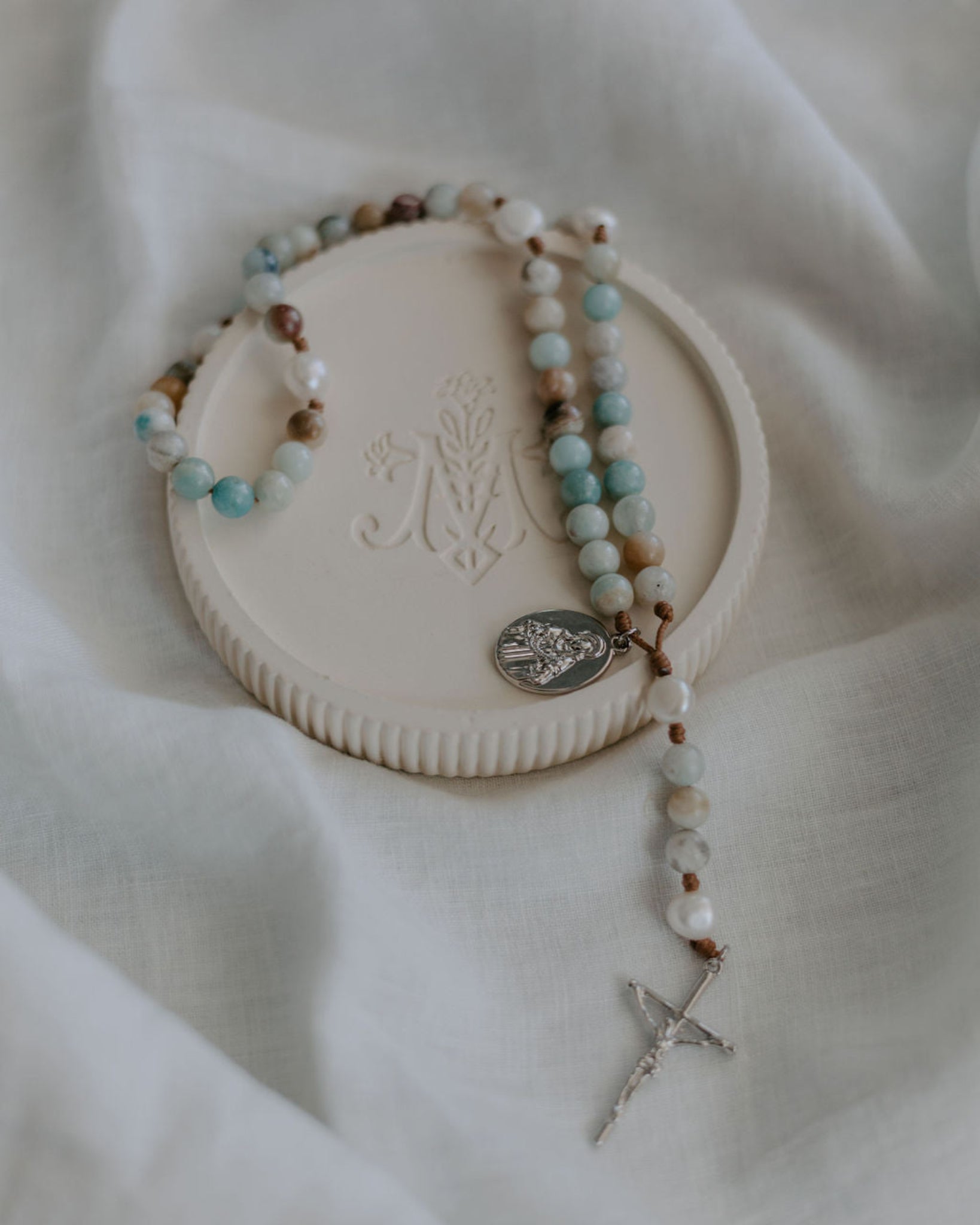 Rosary with turquoise and brown beads on a rosary dish with marian logo, on a white fabric background