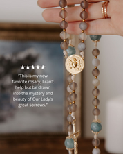 Close-up of a rosary with a gold medal, held by a hand, with a blurred background with 5 star review
