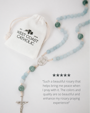 Rosary with blue beads and a silver cross on a white background, accompanied by a drawstring bag labeled 'West Coast Catholic'.