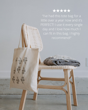 Beige tote bag with floral patterns hanging on a wooden chair against a light gray wall.
