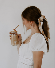 Woman in a white dress holding a jar with a straw against a plain background