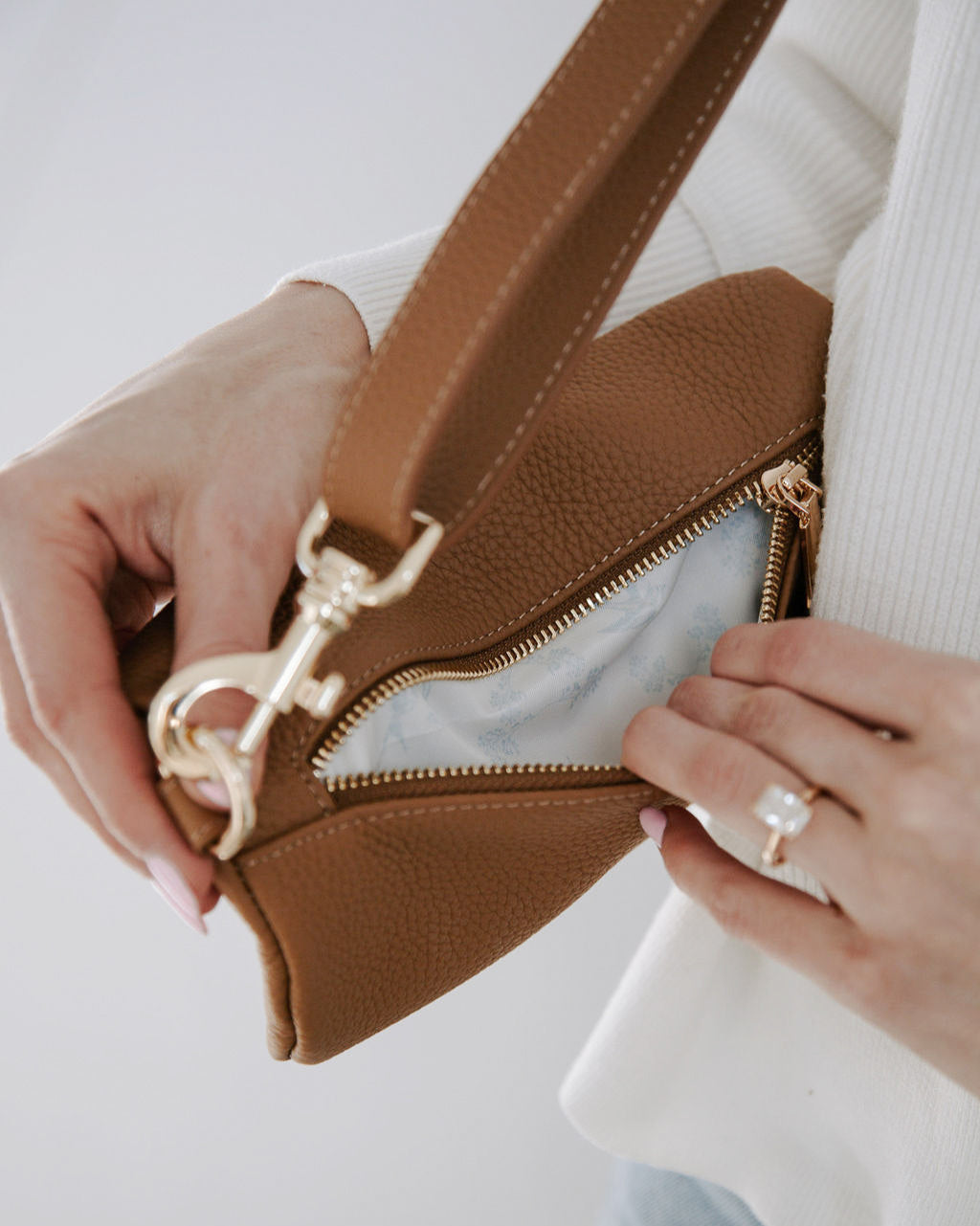 Brown leather handbag with gold zipper being held by a person wearing a white sweater.