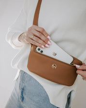 Person holding a brown crossbody bag with a white phone case inside, wearing a white sweater and blue jeans.