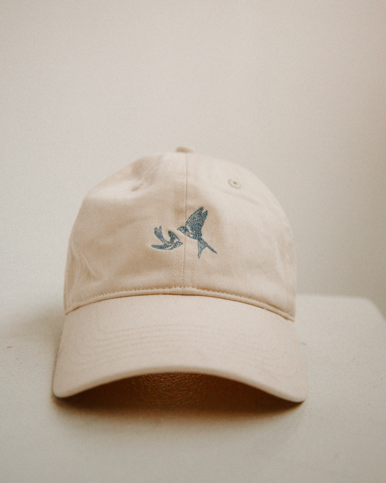 Beige cap with a small embroidered blue swallows design on a light beige background