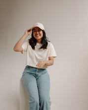 Woman wearing a cream t-shirt and blue jeans, posing with a beige cap against a plain background