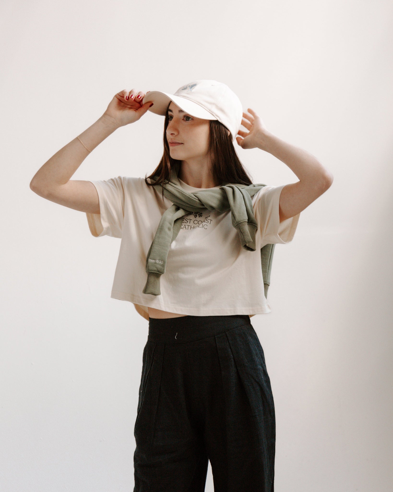 Person wearing a cream crop top with a graphic design, black pants, and a cream hat with blue swallows on a plain background