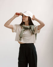 Person wearing a cream crop top with a graphic design, black pants, and a cream hat with blue swallows on a plain background