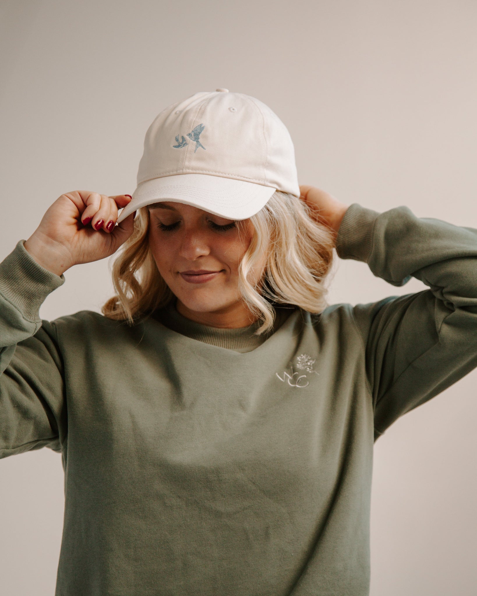 Woman wearing a cream ball cap with blue embroidered swallows and a green crewneck