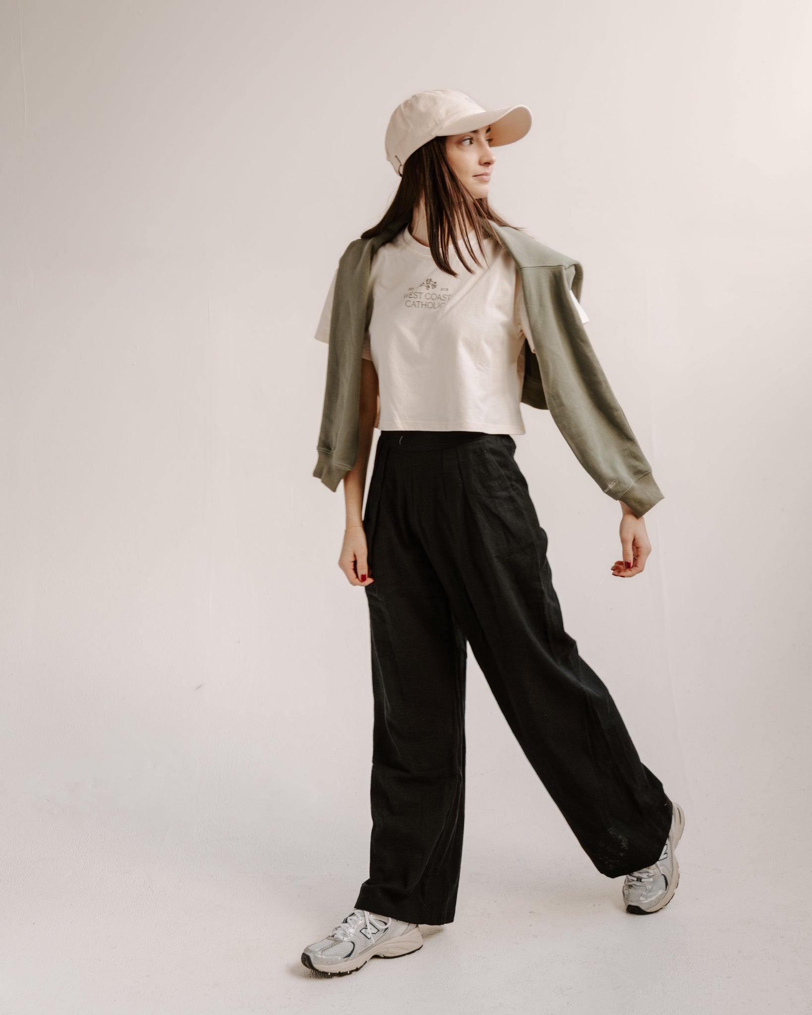 Person wearing a beige shirt, green jacket, black pants, and white sneakers on a white background