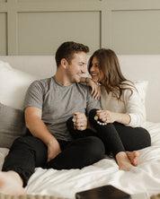 Man and woman sitting on a bed, smiling and holding rosaries and hands.