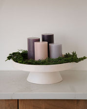 Decorative advent wreath arrangement with candles and greenery on a white pedestal against a neutral background