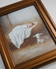 Framed artwork of a baby wrapped in a blanket next to a lamb on a textured surface.