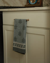 A dark blue kitchen towel with a repeating Marian 'M' pattern and a striped border hanging from a brass cabinet handle in a kitchen setting.