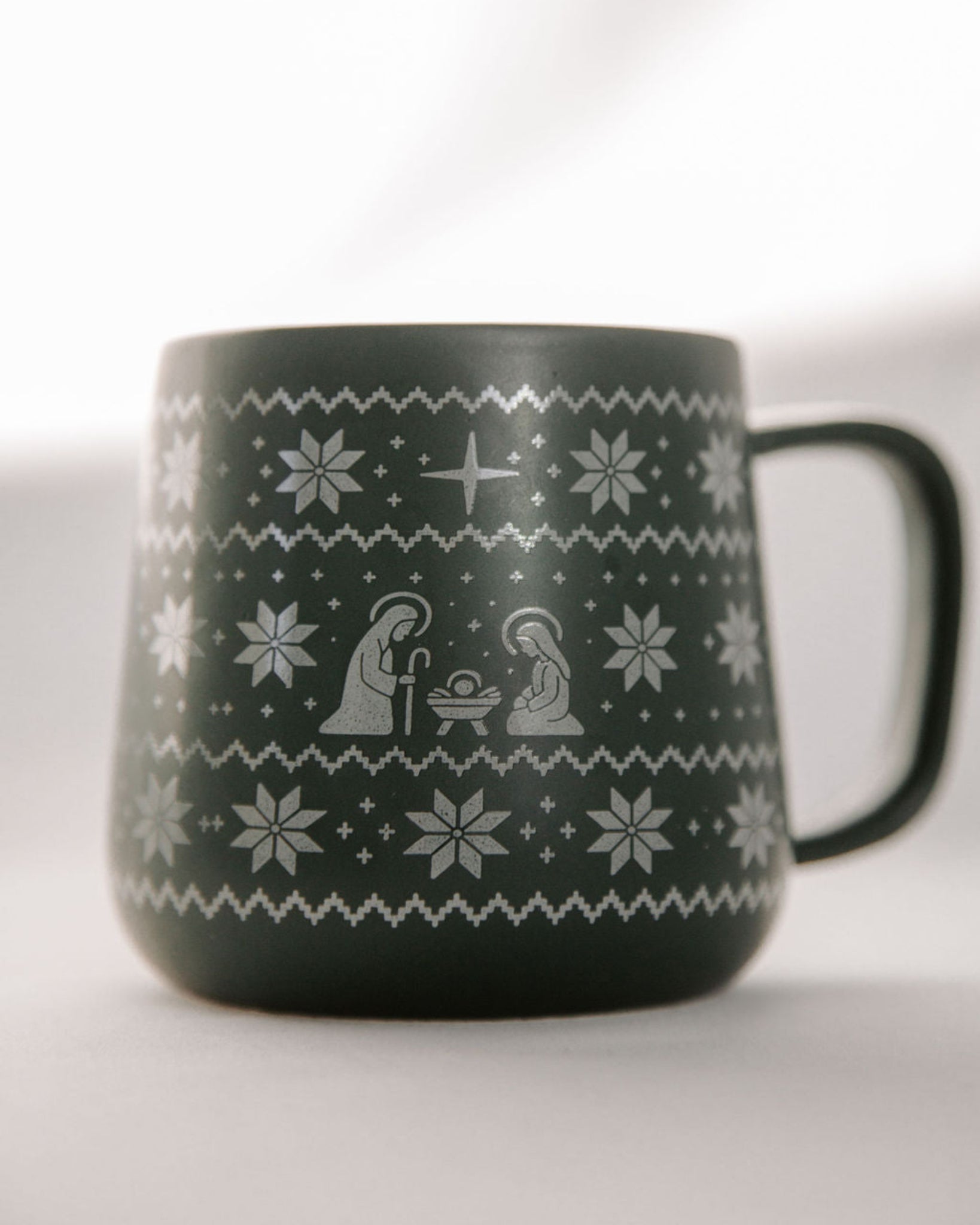Green mug with white nativity scene and snowflake pattern on a white background