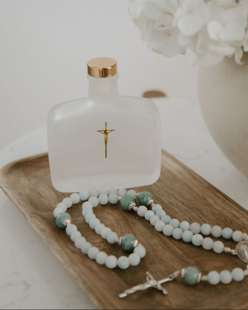 Holy water bottle with gold cap, rosary, and white flowers on a wooden tray