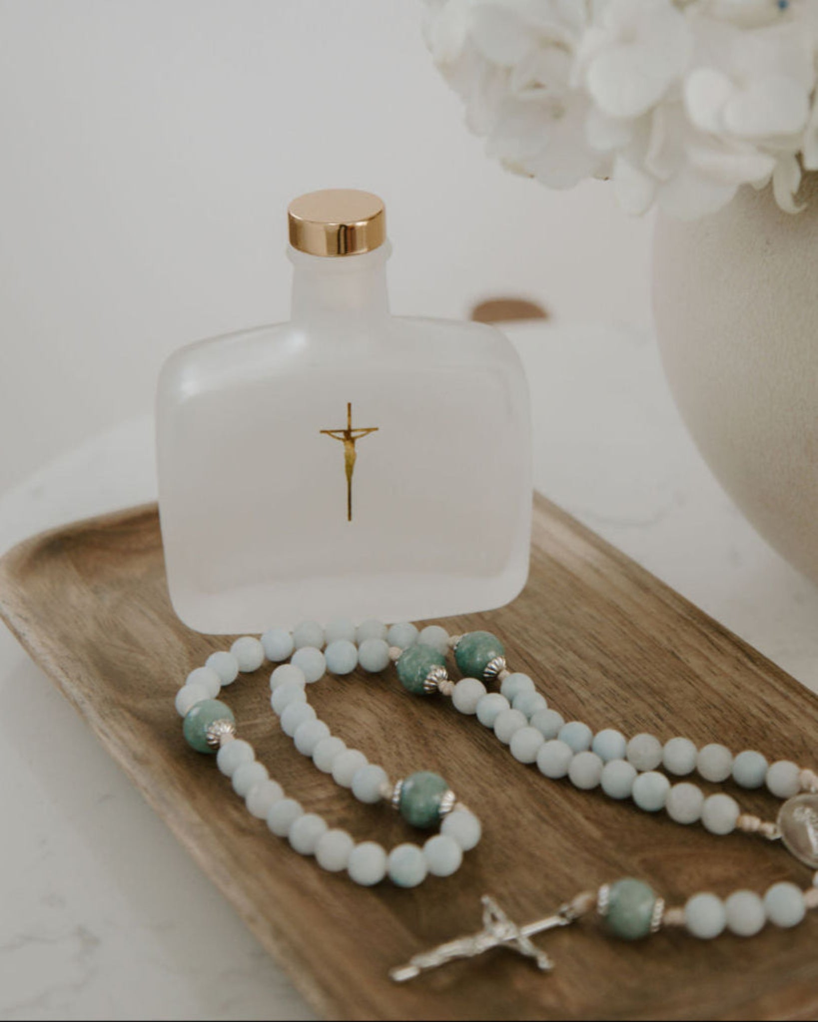 Holy water bottle with gold cap, rosary, and white flowers on a wooden tray