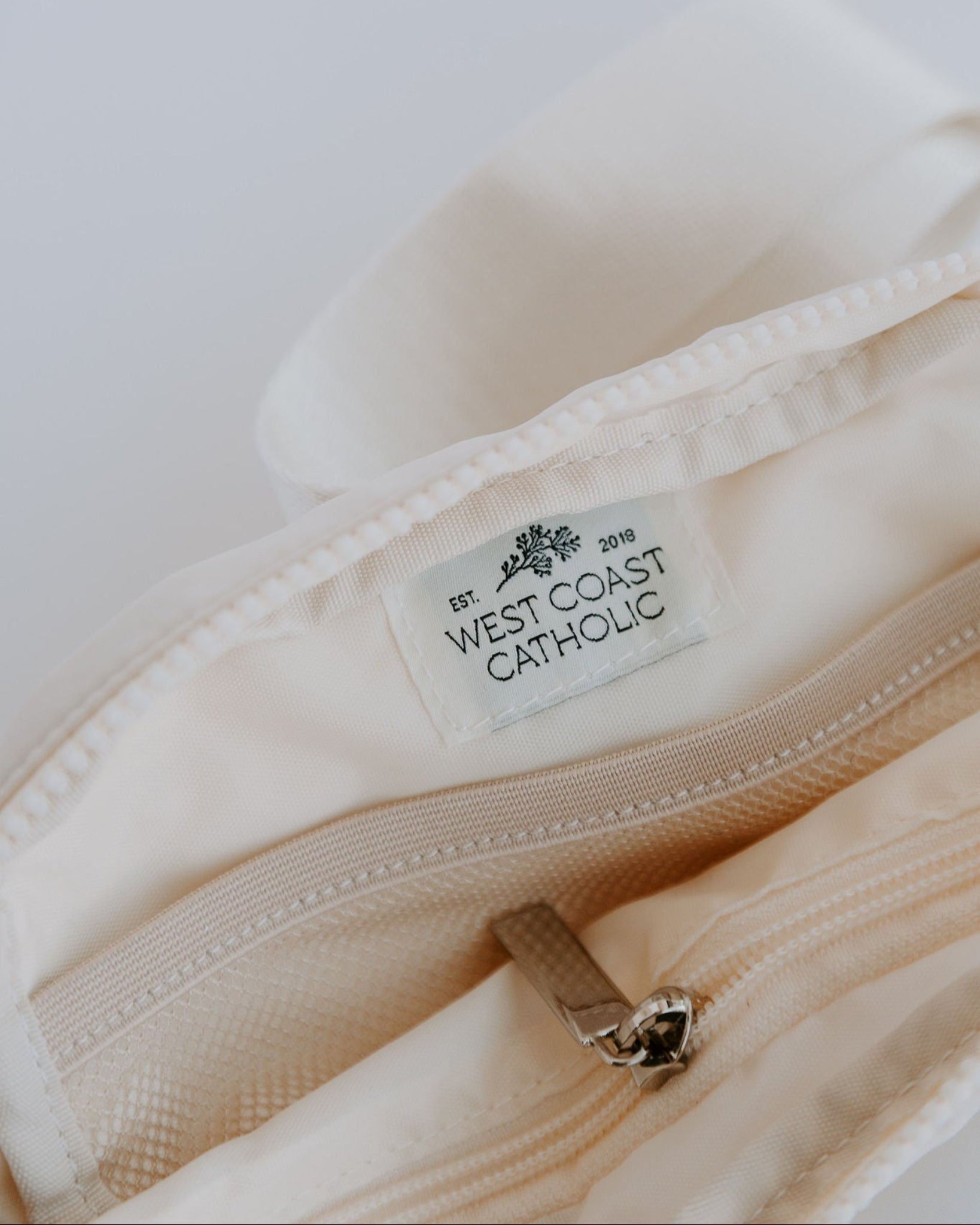 Close-up of a cream marian belt bag with a 'West Coast Catholic' label on a white background