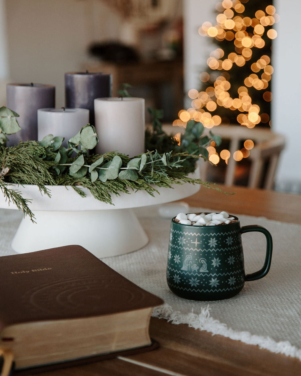 Cozy indoor setting with candles, a mug of hot chocolate, and a book on a table with a blurred Christmas tree in the background.
