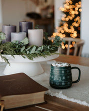 Cozy indoor setting with candles, a mug of hot chocolate, and a book on a table with a blurred Christmas tree in the background.