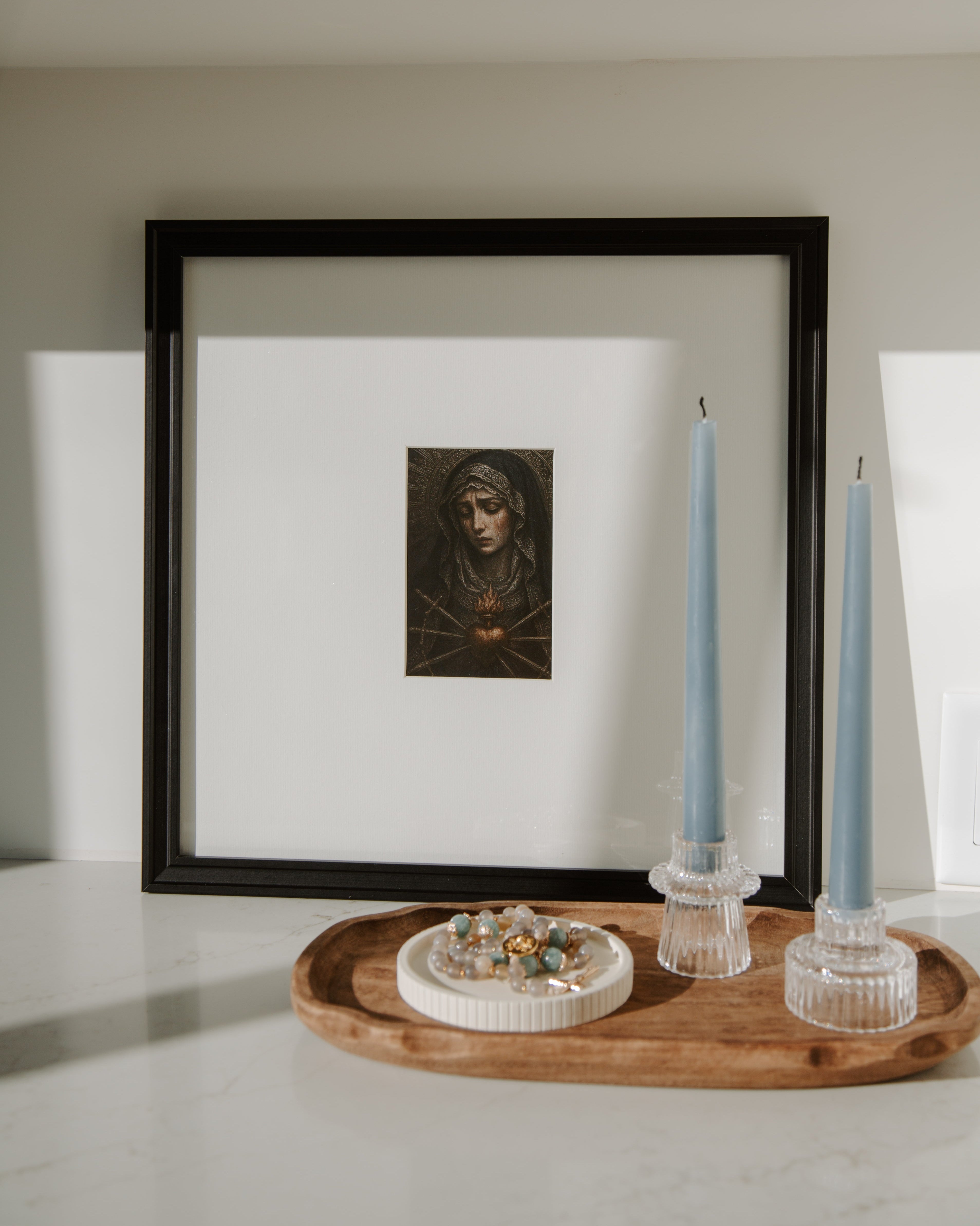Framed artwork of our lady of sorrows a wall with candles and a wooden tray below