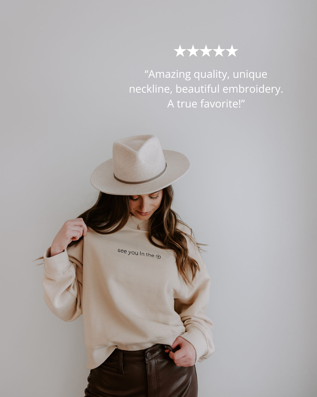 Person wearing a beige sweater with text, holding a beige hat against a plain background.