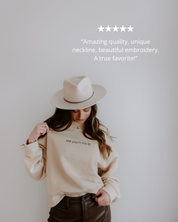 Person wearing a beige sweater with text, holding a beige hat against a plain background.