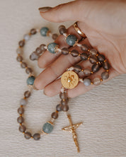 Hand holding rosary with brown and blue beads with gold accents on a white background