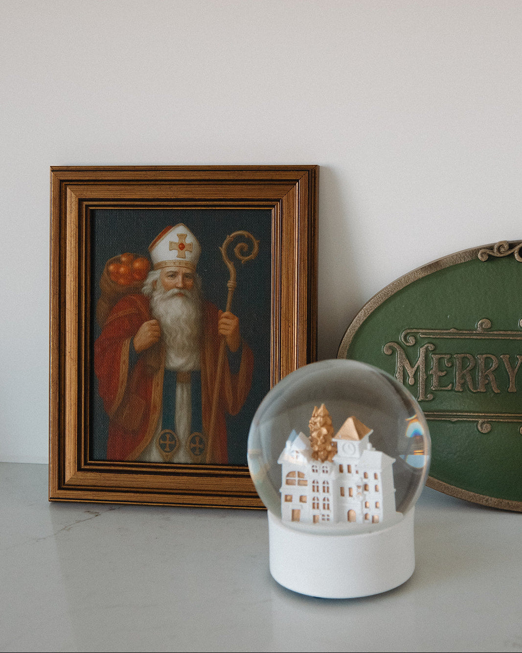 Framed picture of a bearded figure with a staff, snow globe with a castle, and 'Merry' sign on a light surface.