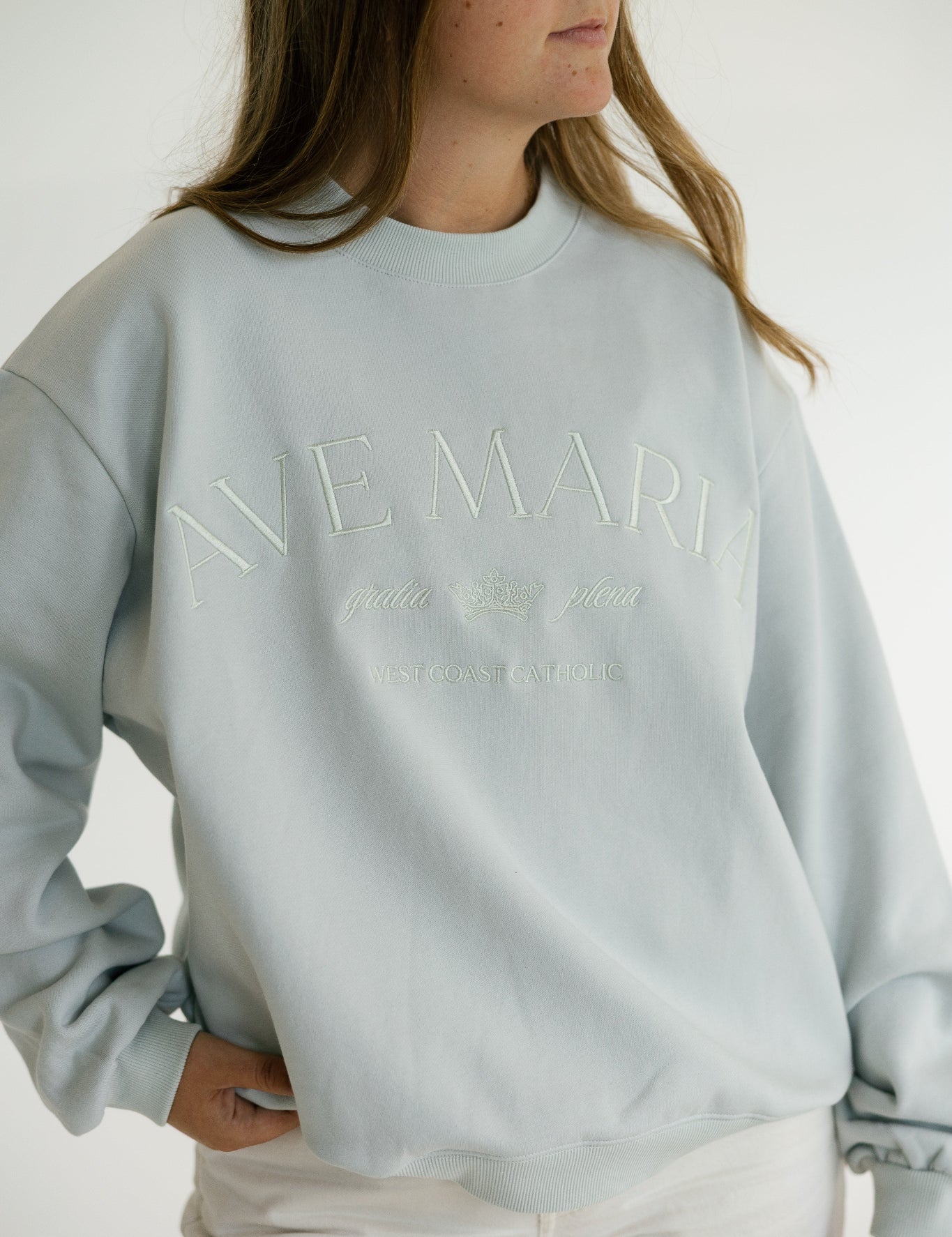 A woman wearing a pale blue crewneck sweatshirt with tonal embroidered text reading 'AVE MARIA' and smaller script underneath 'gratia plena', paired with white pants against a plain light background.