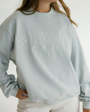 A woman wearing a pale blue crewneck sweatshirt with tonal embroidered text reading 'AVE MARIA' and smaller script underneath 'gratia plena', paired with white pants against a plain light background.