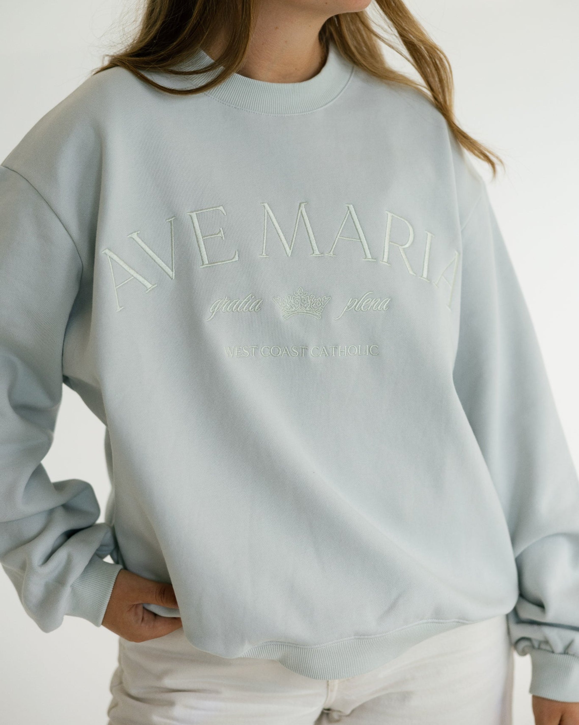 A woman wearing a pale blue crewneck sweatshirt with tonal embroidered text reading 'AVE MARIA' and smaller script underneath 'gratia plena', paired with white pants against a plain light background.