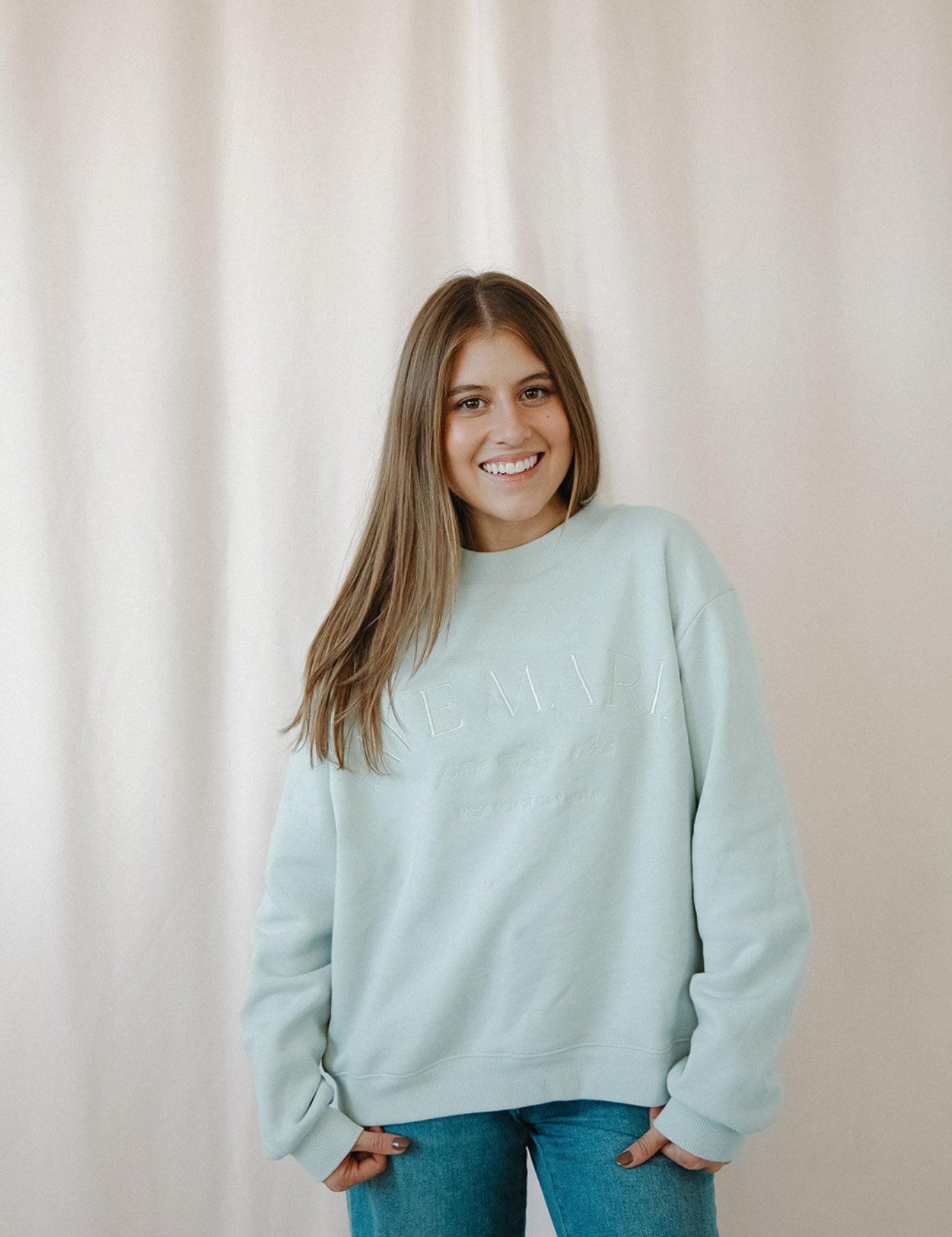 Woman wearing a light blue Ave Maria crewneck and blue jeans against a plain background