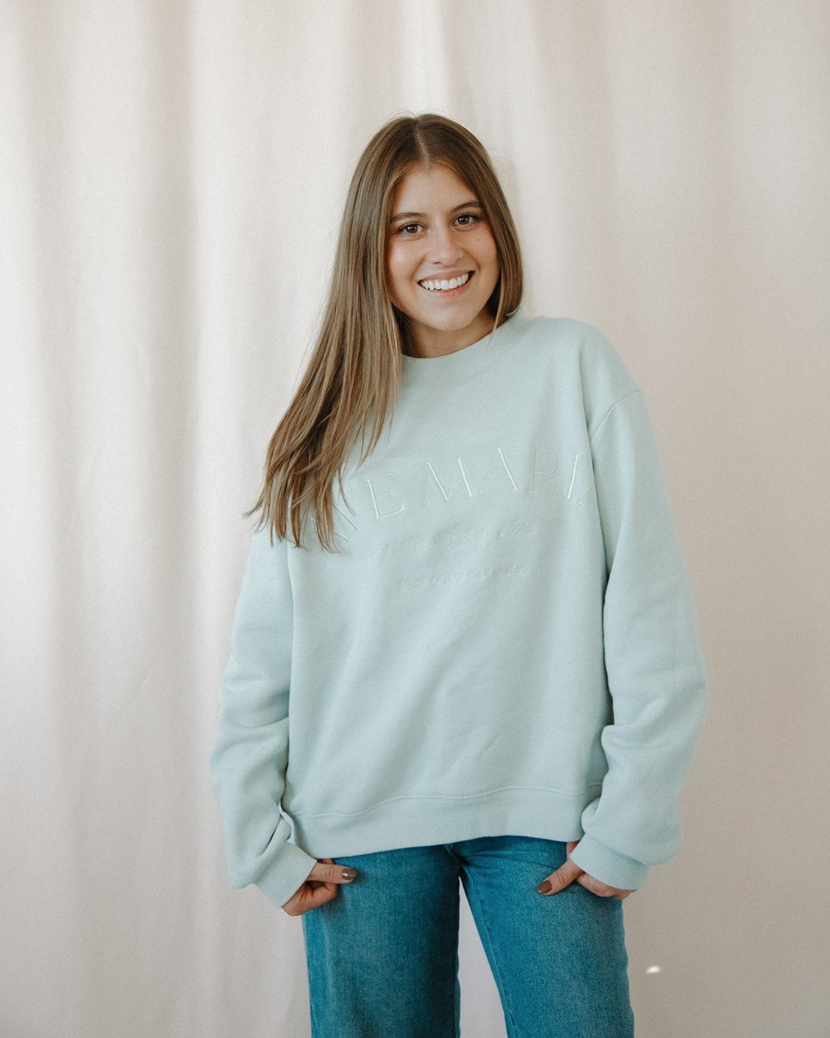 Woman wearing a light blue Ave Maria crewneck and blue jeans against a plain background