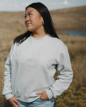 Woman wearing a light gray sweatshirt with Ave Maria text, standing in a scenic outdoor setting.