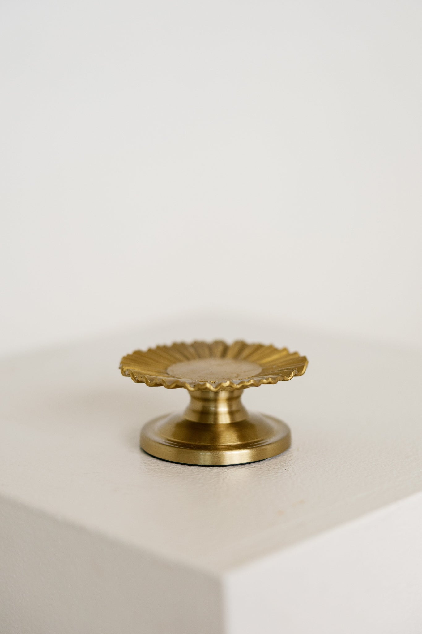 A small brass baptism candle with a scalloped, sunburst-like top and a round base, displayed on a white block against a white background.