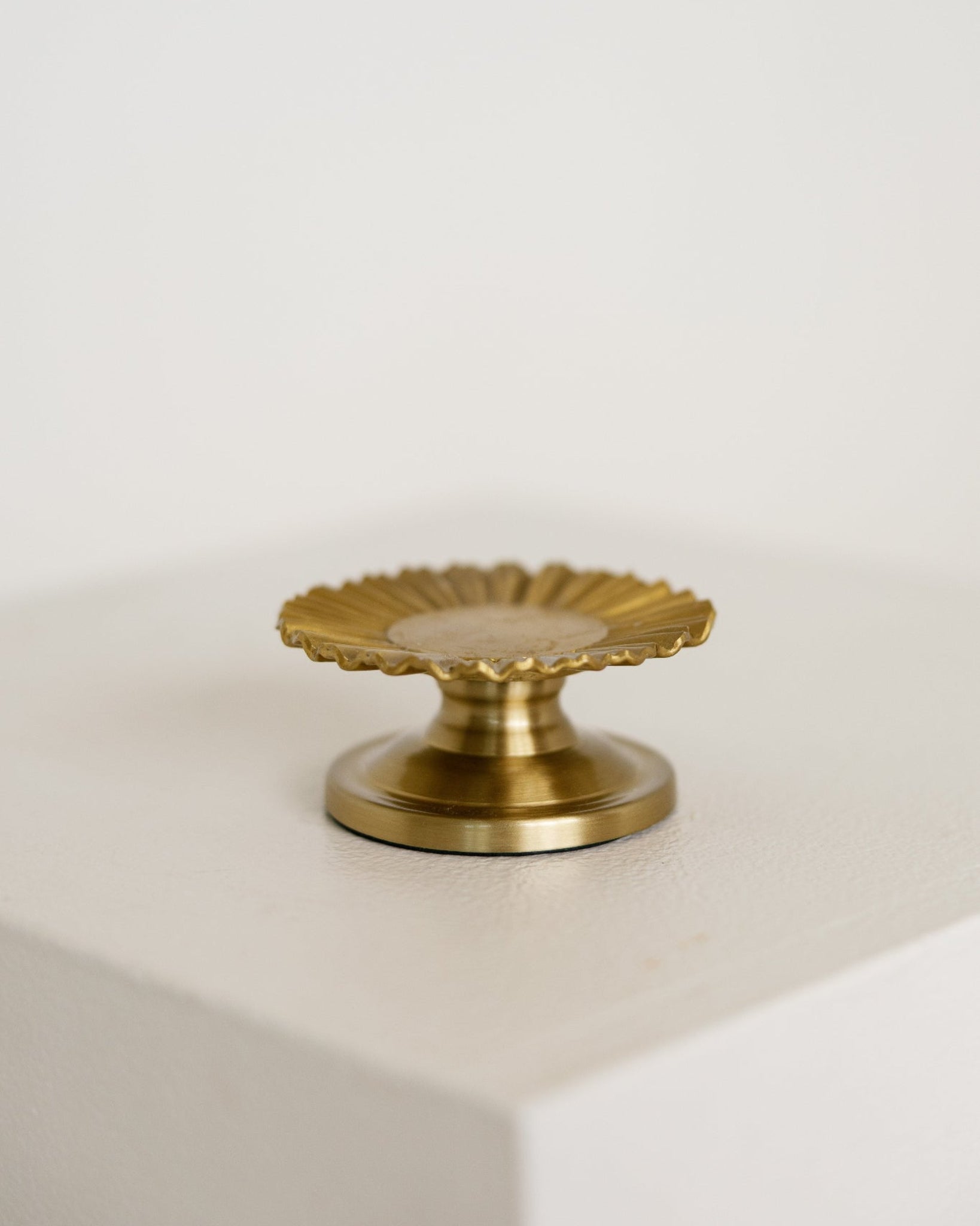 A small brass baptism candle with a scalloped, sunburst-like top and a round base, displayed on a white block against a white background.