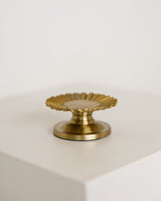 A small brass baptism candle with a scalloped, sunburst-like top and a round base, displayed on a white block against a white background.