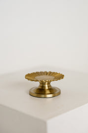 A small brass baptism candle with a scalloped, sunburst-like top and a round base, displayed on a white block against a white background.