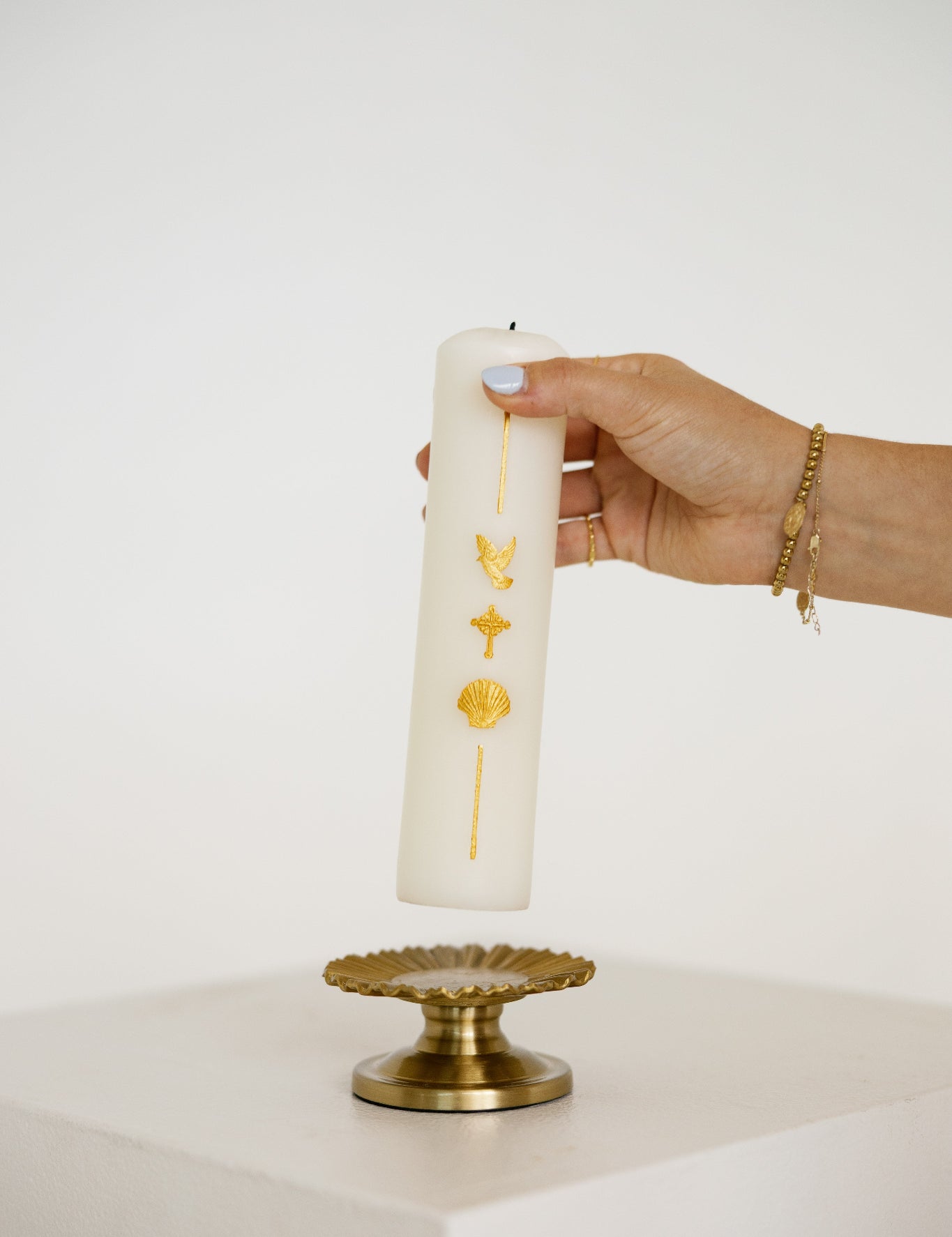 A hand holds a tall white pillar baptism candle decorated with gold symbols, positioned above a gold pedestal against a clean white background.