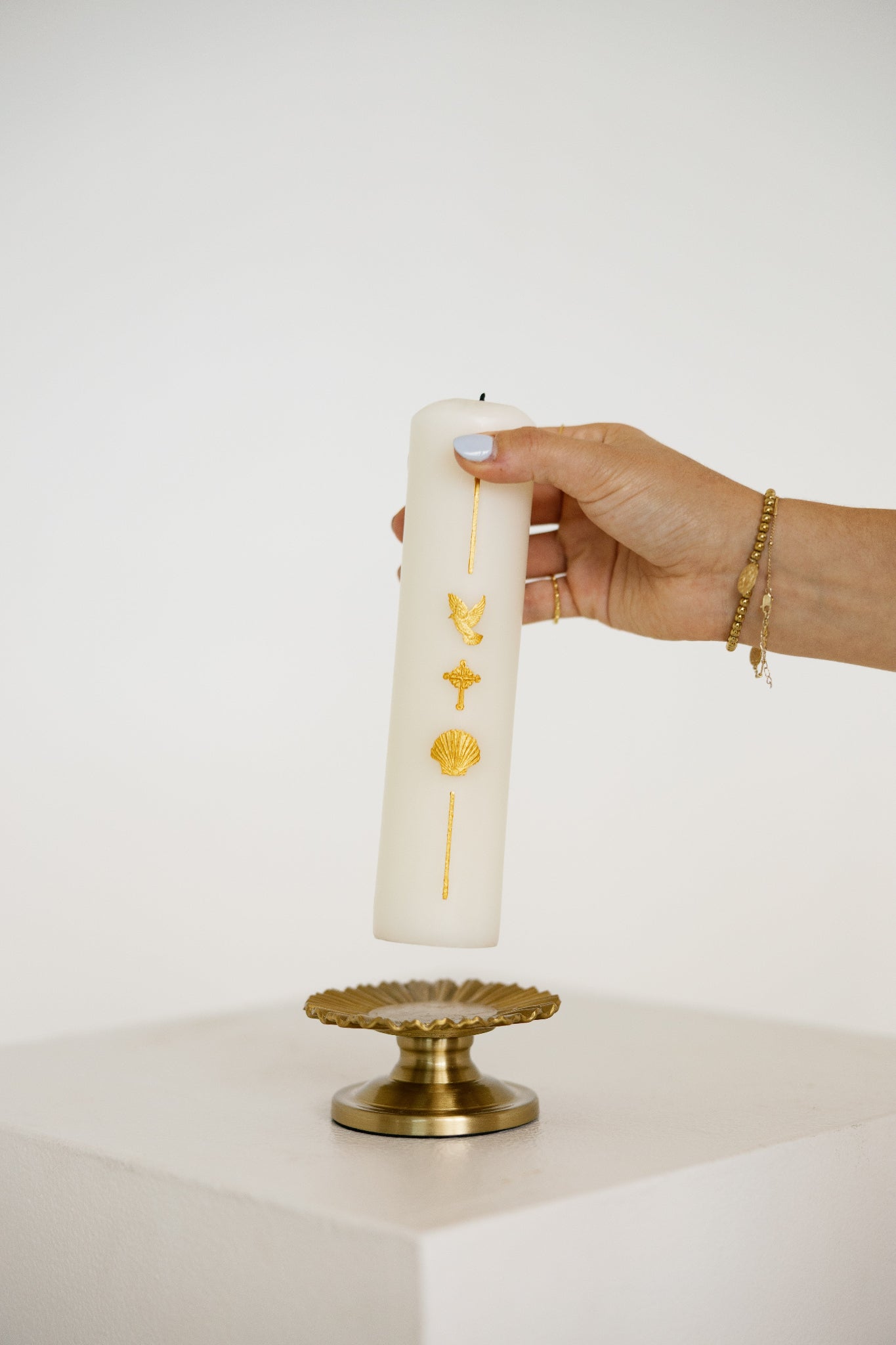 A hand holds a tall white pillar baptism candle decorated with gold symbols, positioned above a gold pedestal against a clean white background.