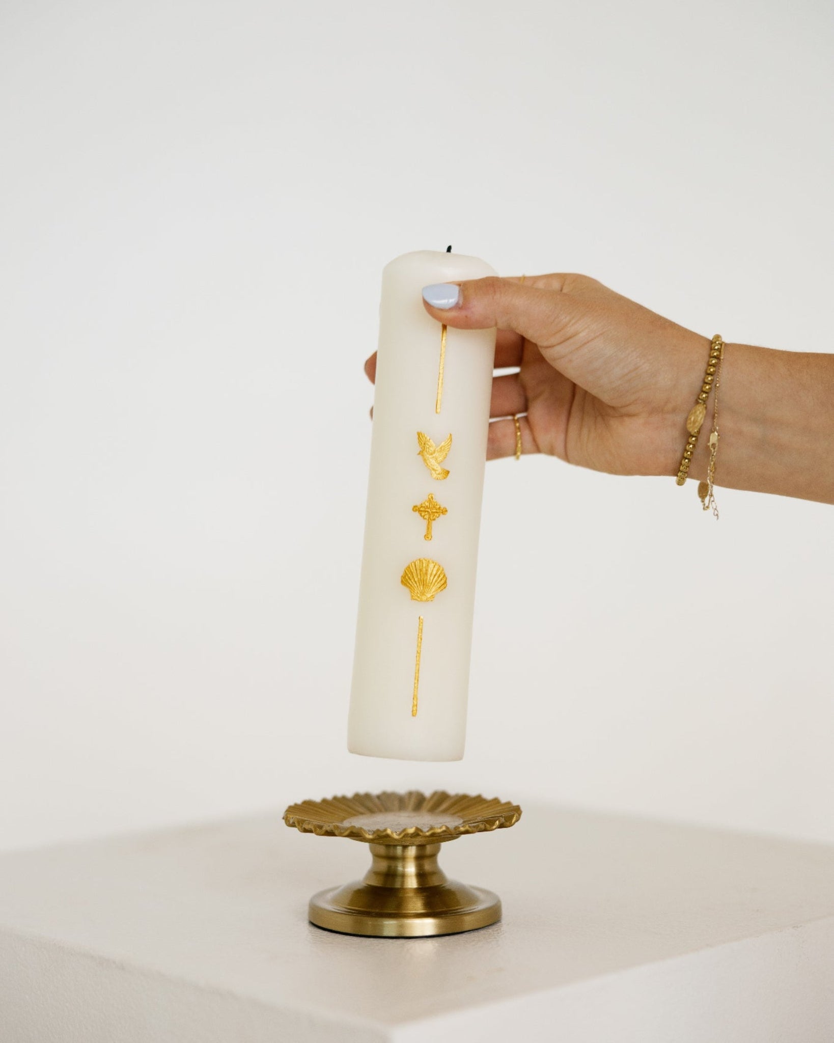 A hand holds a tall white pillar baptism candle decorated with gold symbols, positioned above a gold pedestal against a clean white background.