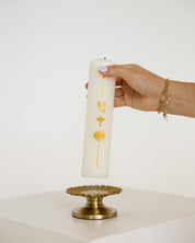 A hand holds a tall white pillar baptism candle decorated with gold symbols, positioned above a gold pedestal against a clean white background.