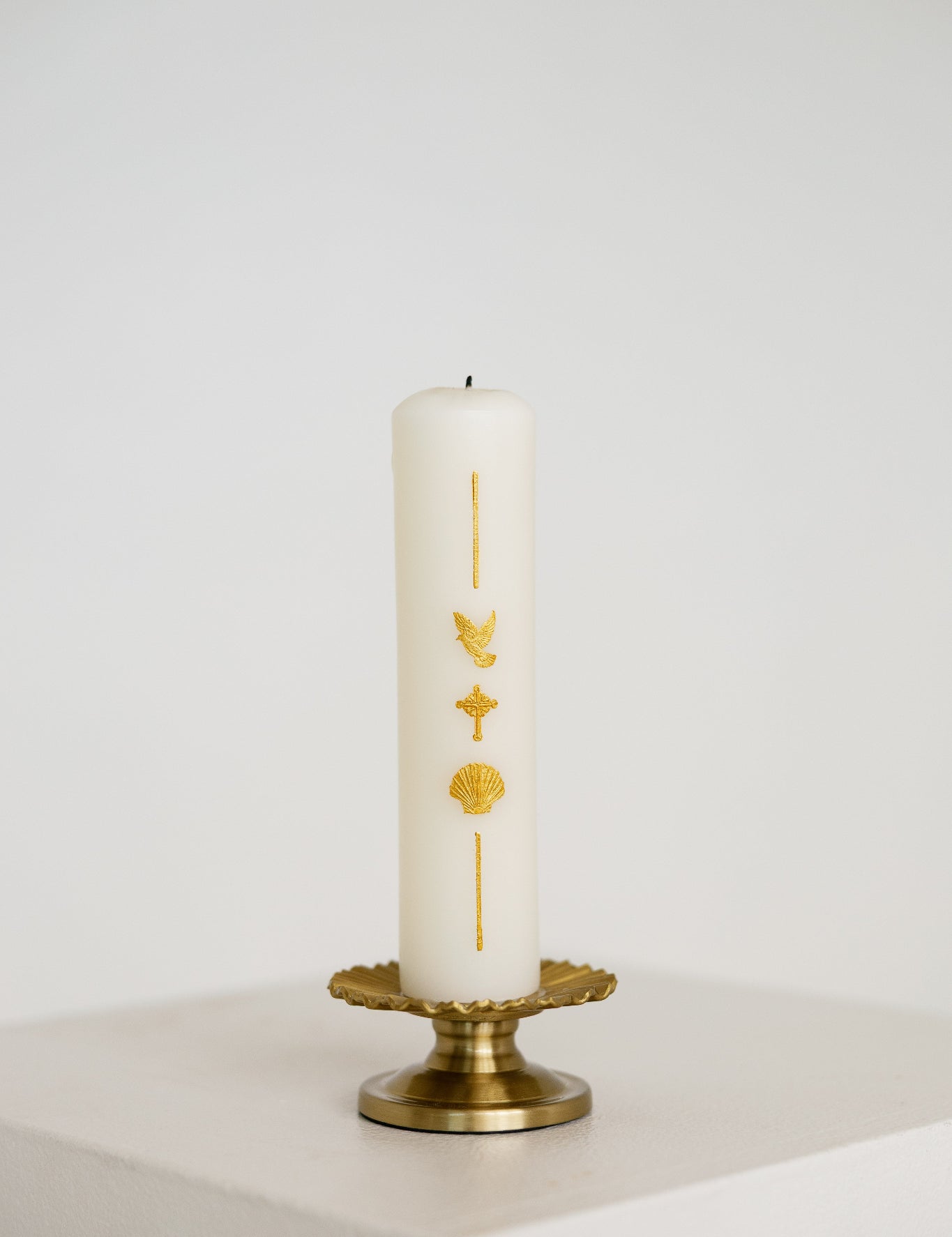 A white pillar Baptism candle with gold religious symbols, displayed on a gold metal candle holder against a clean white background.