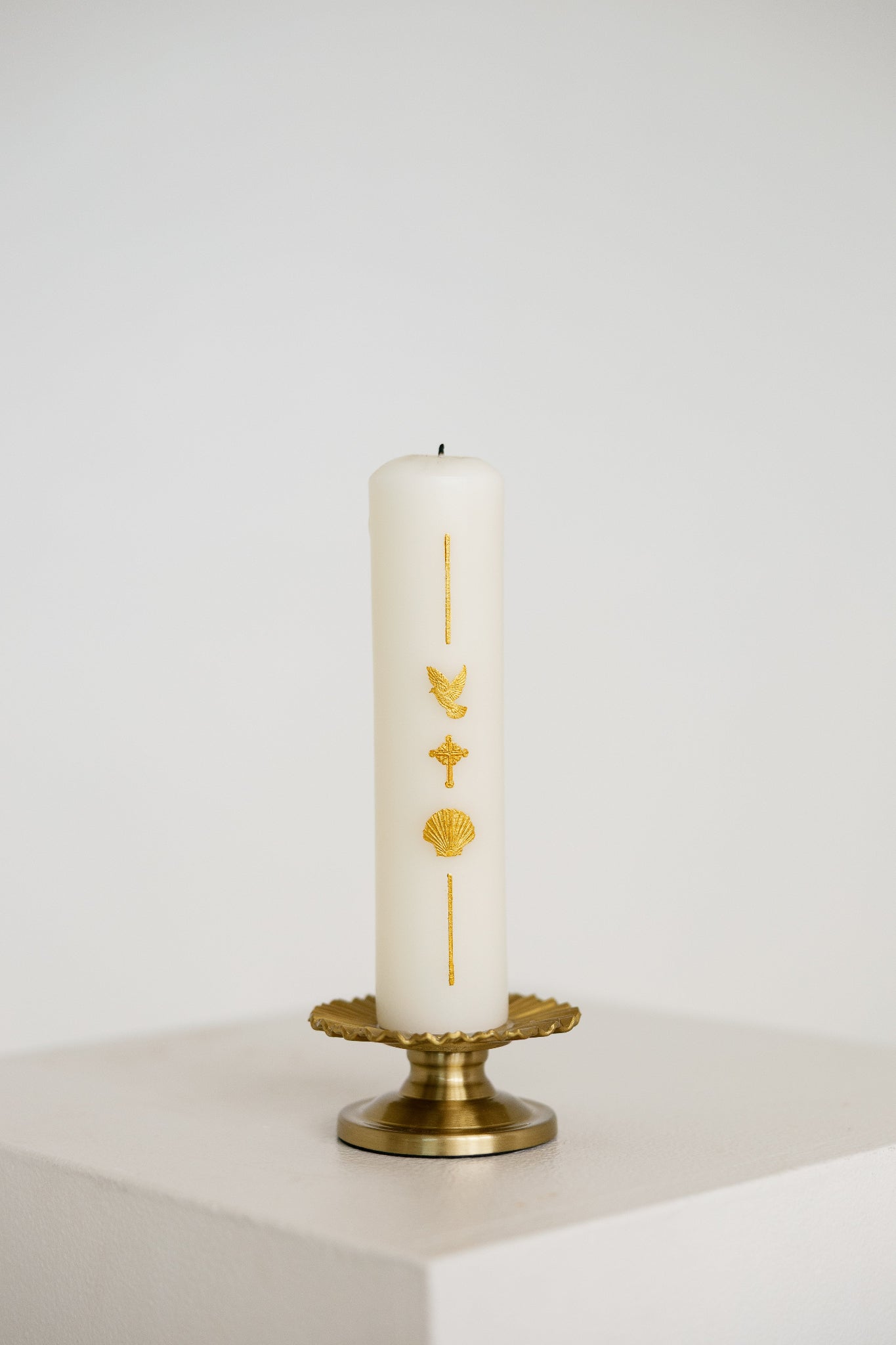 A white pillar Baptism candle with gold religious symbols, displayed on a gold metal candle holder against a clean white background.