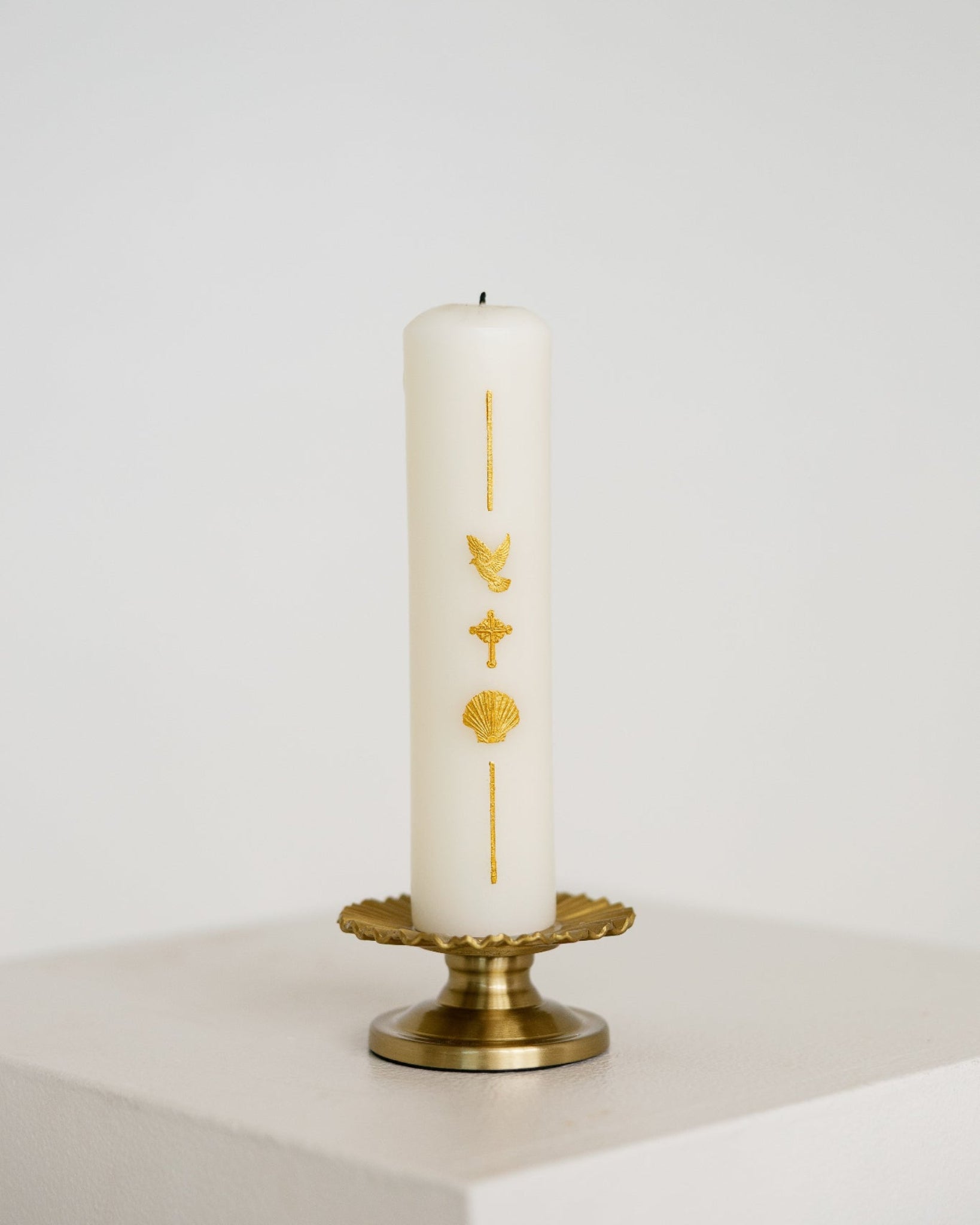 A white pillar Baptism candle with gold religious symbols, displayed on a gold metal candle holder against a clean white background.