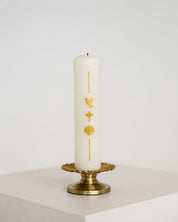 A white pillar Baptism candle with gold religious symbols, displayed on a gold metal candle holder against a clean white background.