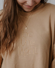 Person wearing a beige sweater with 'be the light' text, smiling.