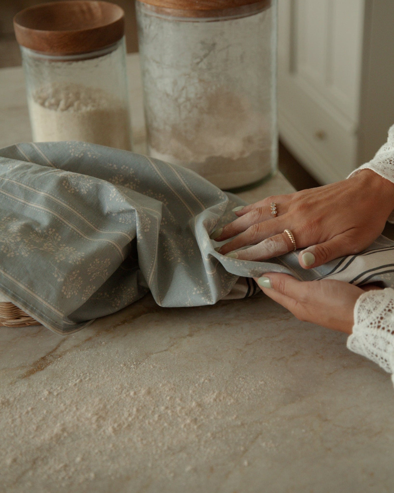 A close-up of a person's hands folding a dusty blue marian linen tea towel with gold stripes on a marble countertop. In the background, glass canisters with wooden lids are visible.