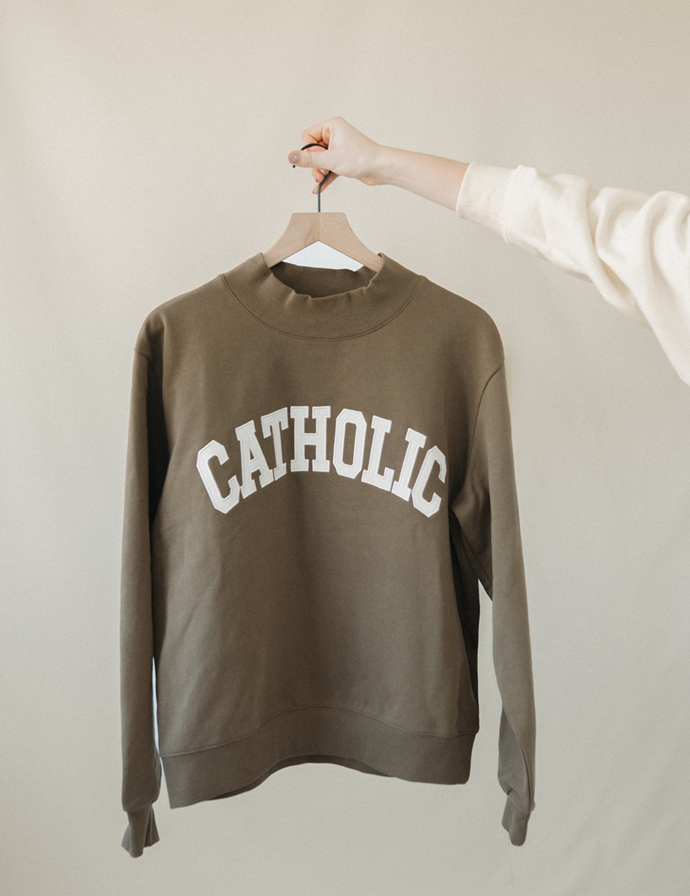 Green sweatshirt with 'CATHOLIC' text on a hanger against a beige background